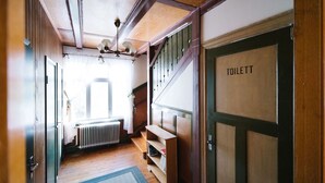 A narrow hallway with wooden floors, a radiator, and a staircase leading to an upper level. A door labeled "TOILETT" is visible on the right. - Urshult Hotell (Urshult)
