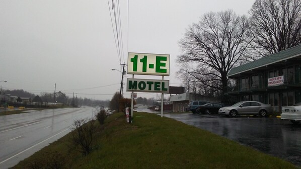 A motel sign with a green '11-E' and 'MOTEL' text, located beside a road with parked cars and a building in the background. - Eleven E Motel (Johnson City)