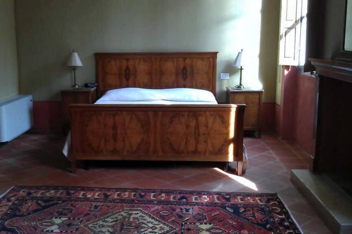 Un dormitorio con una cama de madera, dos mesitas de noche con lámparas, un radiador y una alfombra estampada.