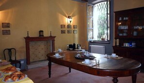 Uma sala de jantar com uma grande mesa de madeira posta para o café da manhã, uma lareira e uma vista pela janela com um portão de metal.