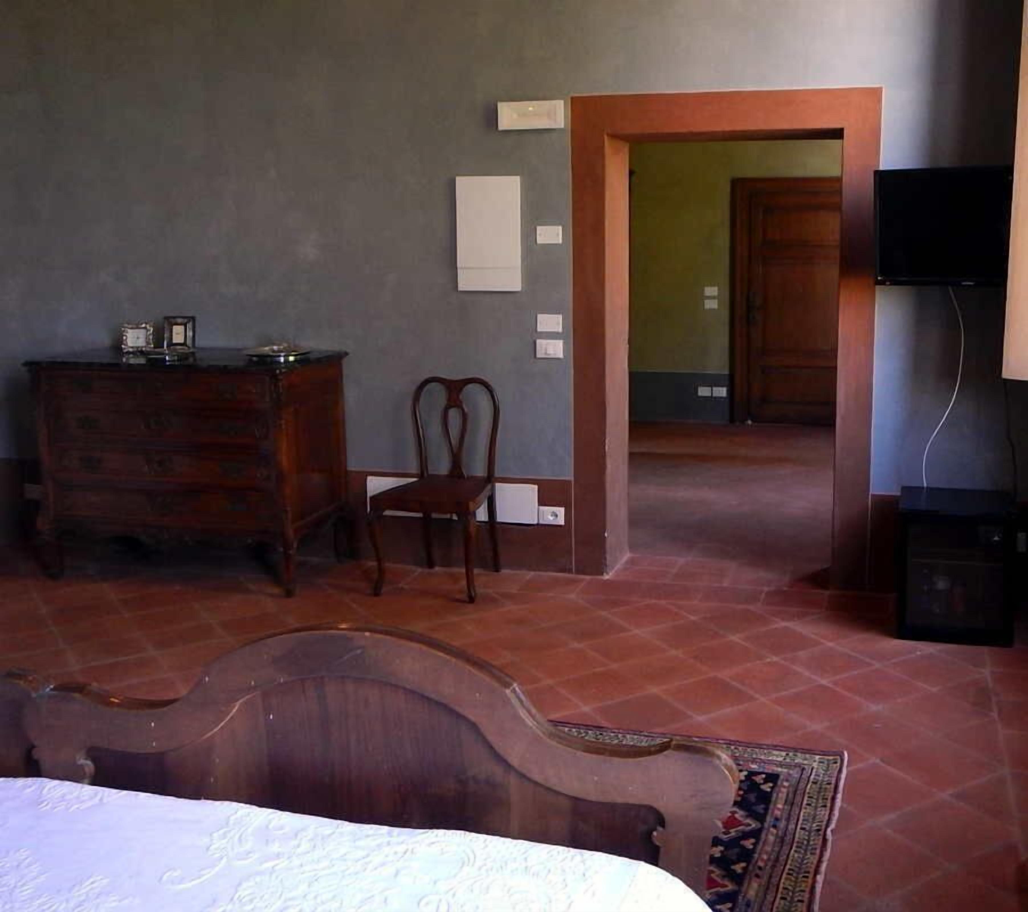 Un dormitorio con un aparador de madera, una silla, una cama con cabecera, un televisor y una puerta que da a otra habitación.