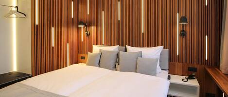 Una habitación de hotel moderna con una cama grande, panel de madera en la pared, mesitas de noche y lámparas fijadas a la pared.