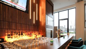 Un área de bar moderna con un televisor montado en la pared mostrando un noticiero, diversas botellas y vasos sobre el mostrador, y vistas de la ciudad a través de amplias ventanas.