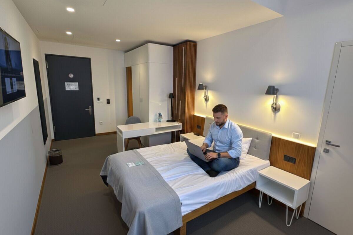 A modern hotel room with a bed, a desk, and a chair. There is a door and a wall-mounted light fixture.