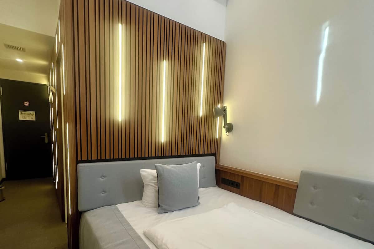 A hotel room with a bed, a headboard with vertical wooden slats, a wall-mounted light fixture, and a door with a sign.