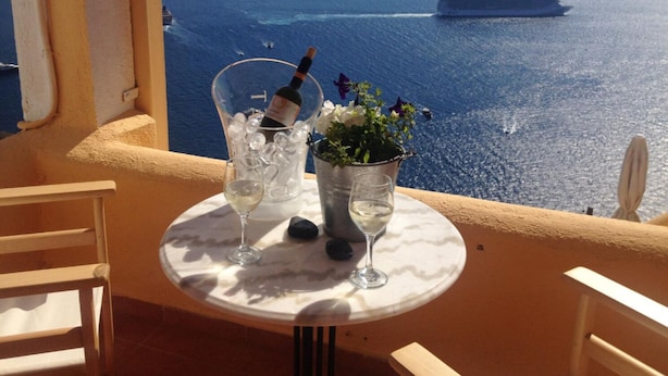 A table with two glasses of white wine, a bottle of wine in a bucket, and a vase with flowers, overlooking a scenic view of the sea and islands. - Hotel Porto Carra (Santorini)