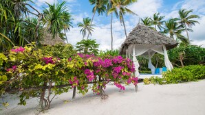 A beach scene with a thatched-roof gazebo, palm trees, and vibrant bougainvillea flowers. - Zanzibar Queen Hotel (Matemwe)