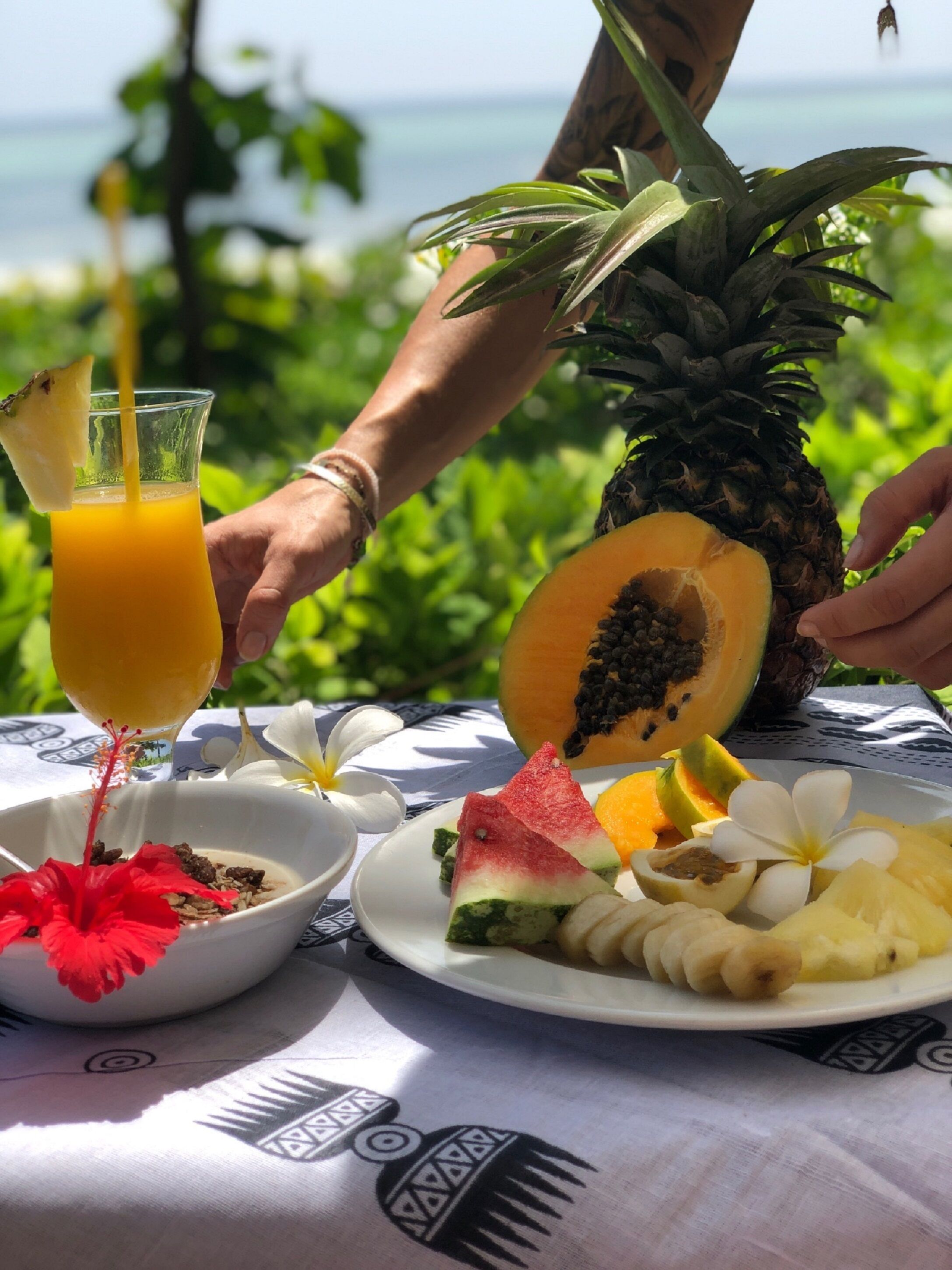 A tropical breakfast setting with a pineapple, papaya, and various fruits on a table, with a view of the ocean in the background.