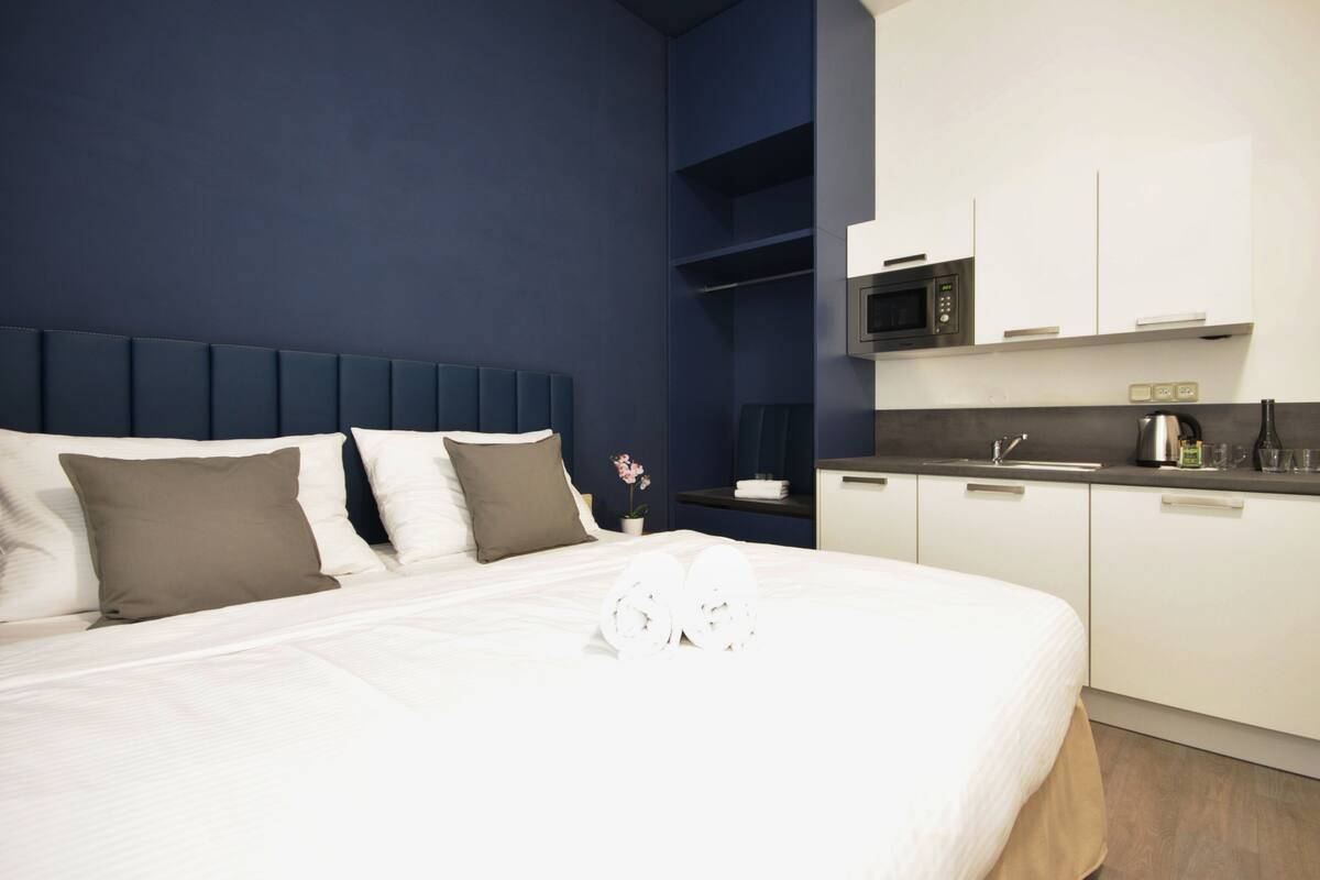 Habitación de hotel con una cama grande, un cabecero azul oscuro, una mesita de noche blanca con una planta, un microondas y una pequeña cocina con una tetera.