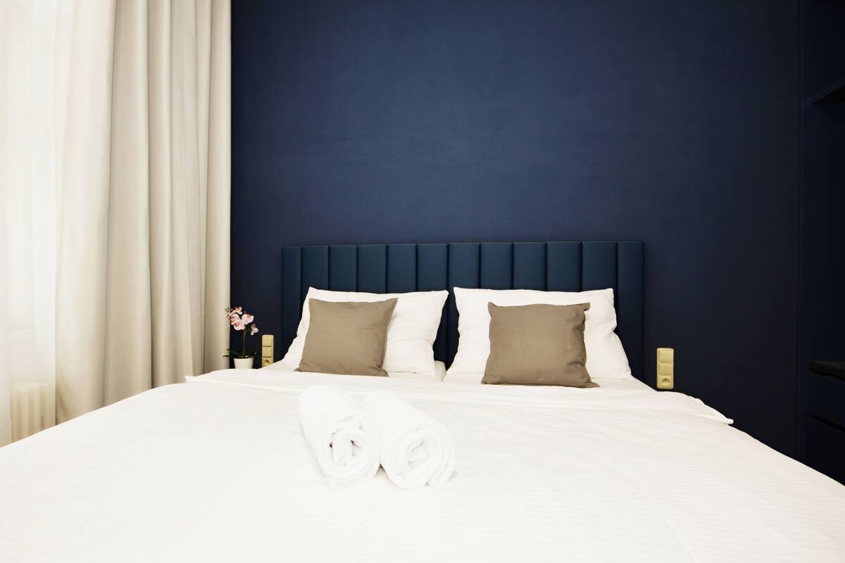 Una cama bien hecha con sábanas blancas y dos almohadas, un cabecero azul oscuro y una cortina blanca a la izquierda.