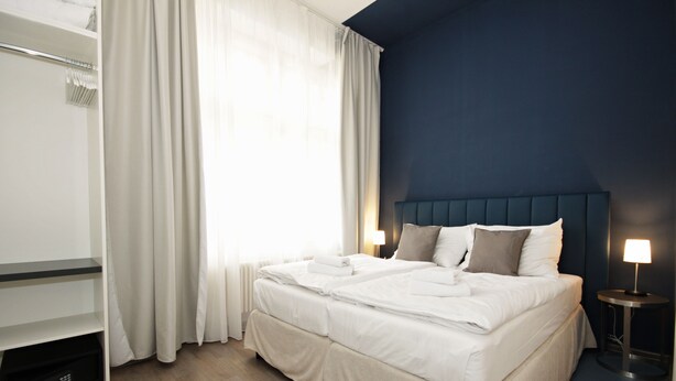Een hotelkamer met een tweepersoonsbed, een kledingkast, een nachtkastje met een lamp en een groot raam met gordijnen.