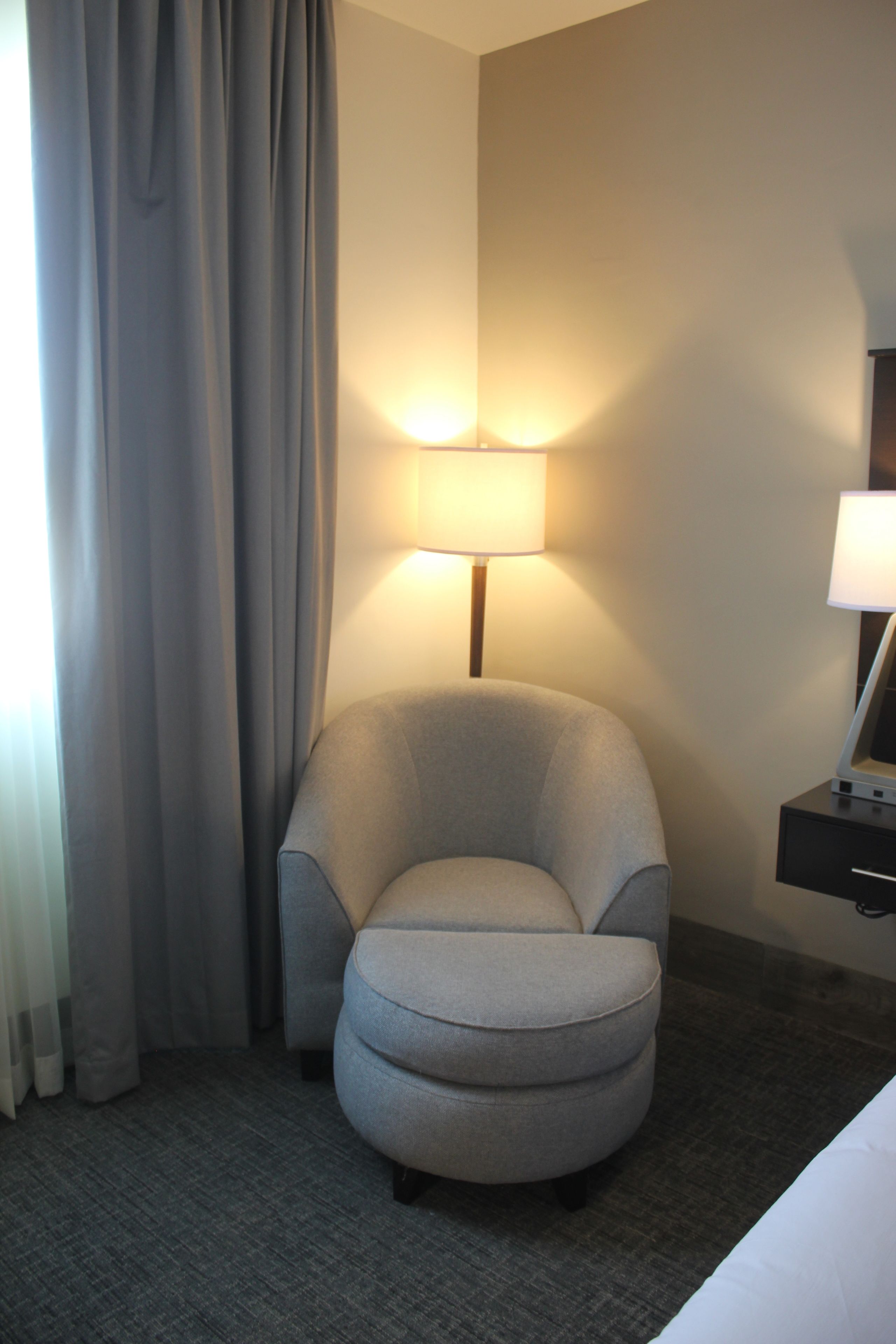 A hotel room with a grey armchair, a lamp, a bed, and a nightstand.