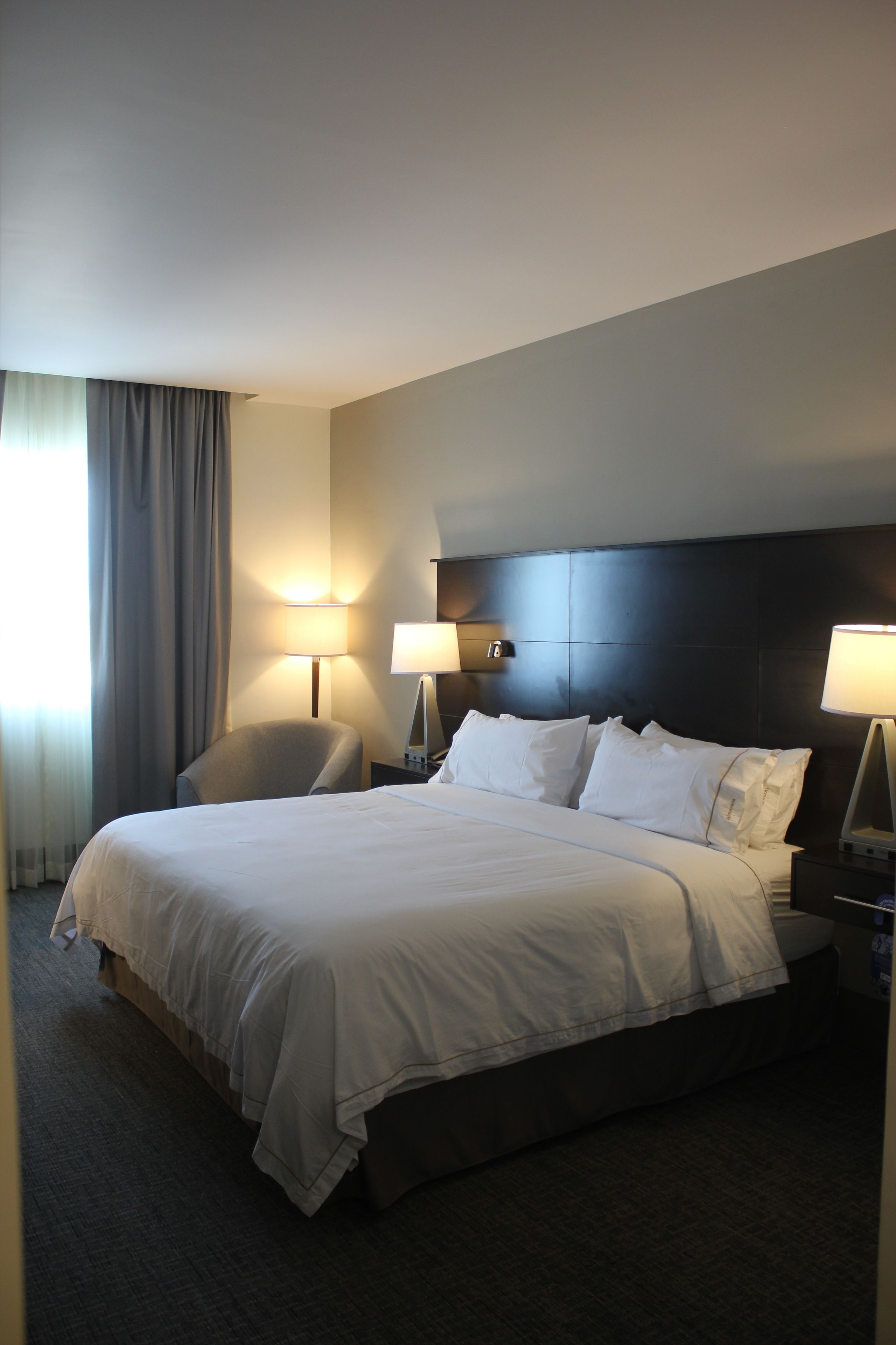 A hotel room with a large bed, two bedside lamps, a chair, and a window with curtains.