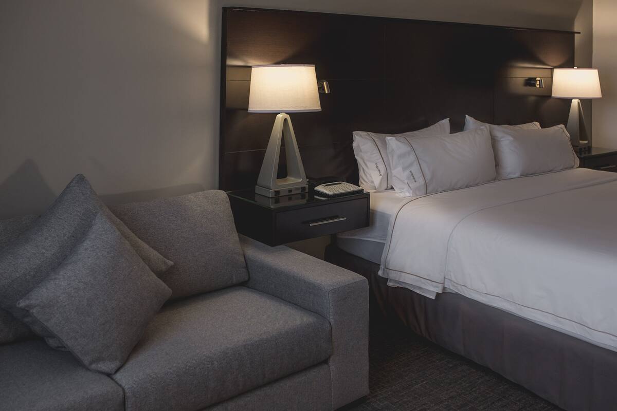 Een hotelkamer met een bed, een nachtkastje met een lamp, een kleine tafel met een telefoon en een grijze fauteuil.