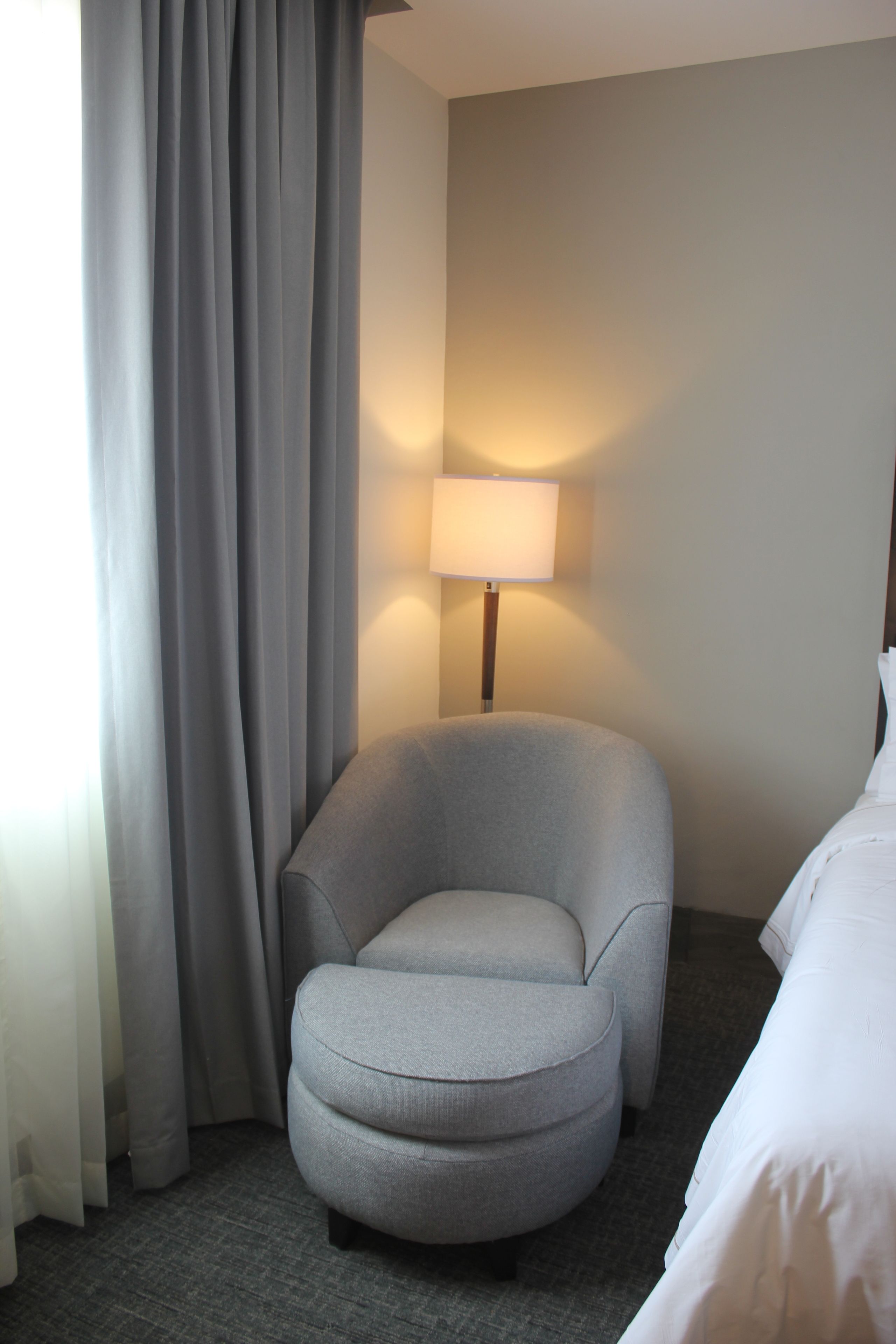 A hotel room with a grey armchair, a bedside table with a lamp, and a bed with white linens.