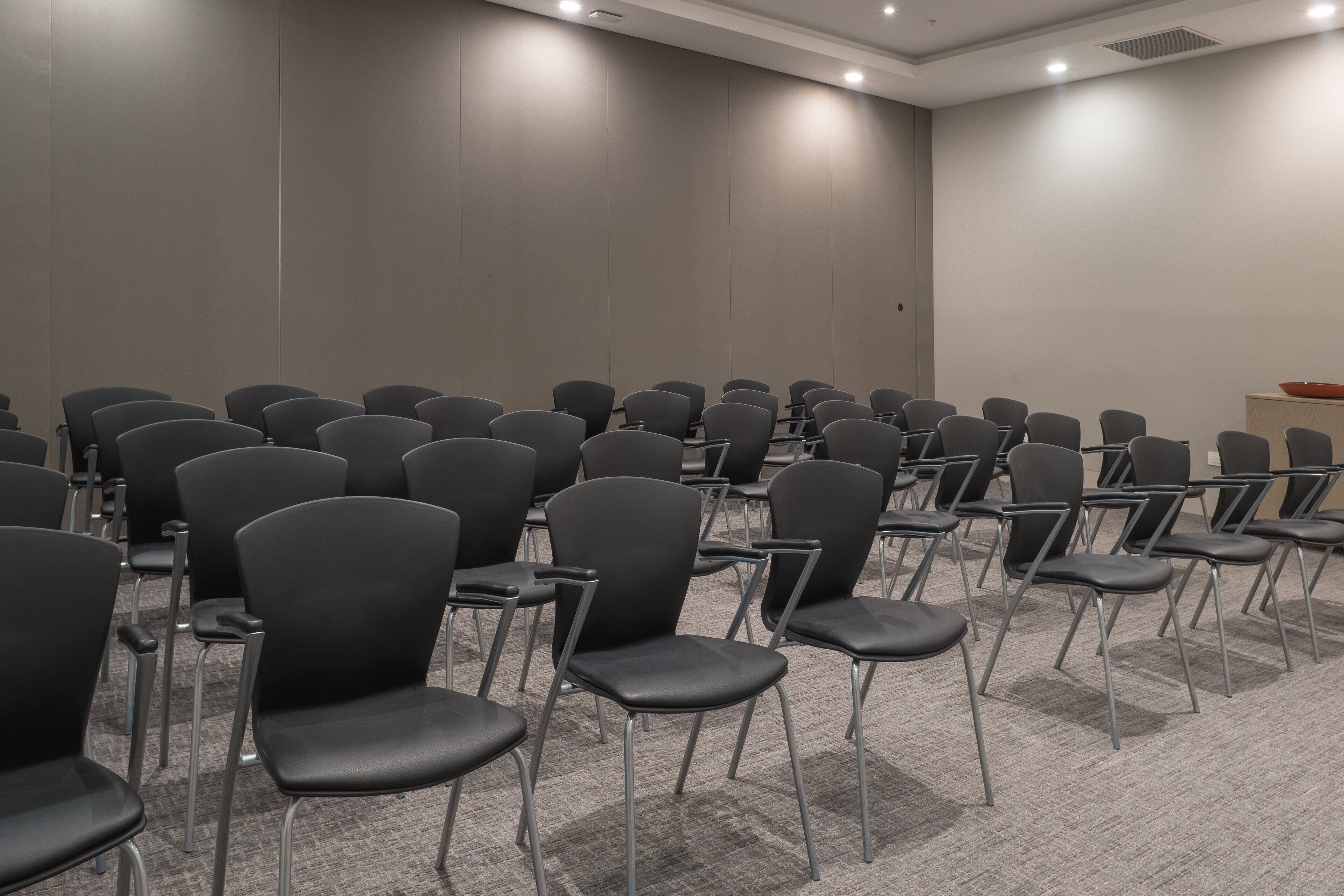 A conference room with rows of black chairs, carpeted floor, and a neutral-colored wall.