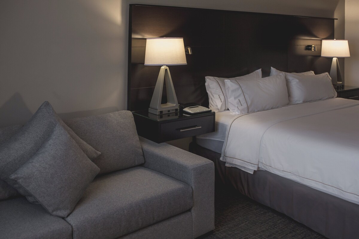 Une chambre d’hôtel avec un lit, une table de chevet avec une lampe, une petite table avec un téléphone et un fauteuil gris.