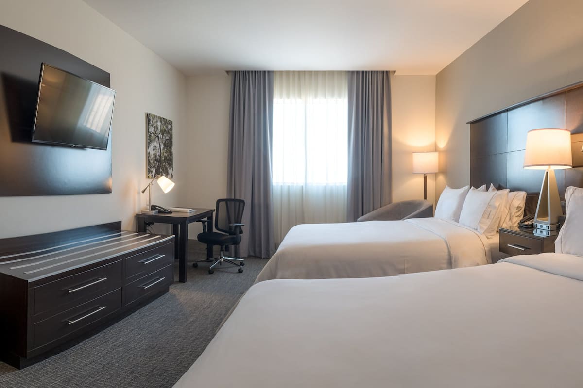 Een hotelkamer met twee bedden, een bureau met stoel, een aan de muur bevestigde televisie en een raam met gordijnen.