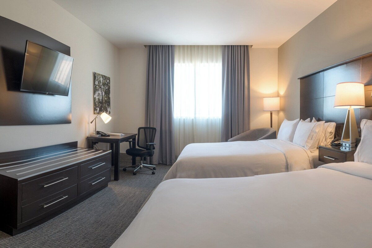 Une chambre d’hôtel avec deux lits, un bureau avec une chaise, un téléviseur fixé au mur et une fenêtre avec des rideaux.