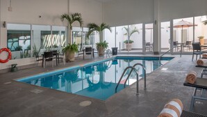 Une piscine intérieure aux parois en verre transparent, entourée de chaises longues et de plantes en pot.