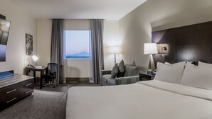 Une chambre d’hôtel avec un lit, un bureau et une chaise, une table de chevet avec une lampe, et une vue sur l’extérieur par une fenêtre.