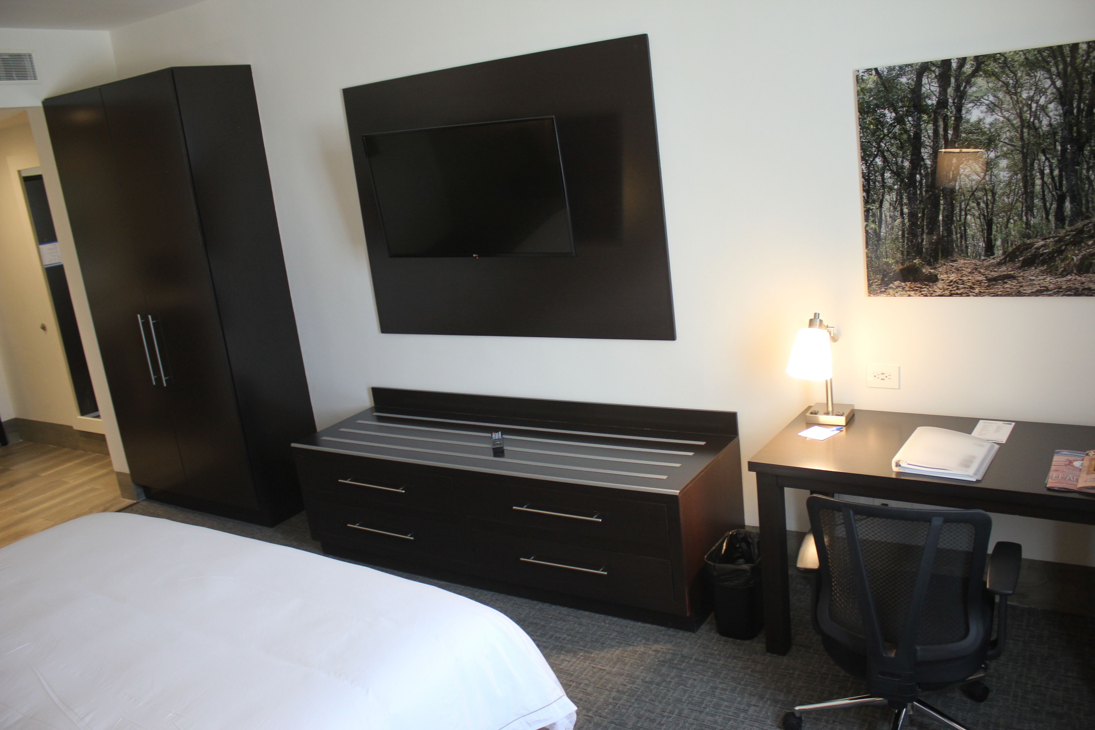 Quarto de hotel com cama, escrivaninha com cadeira, televisão na parede e pintura de uma floresta.