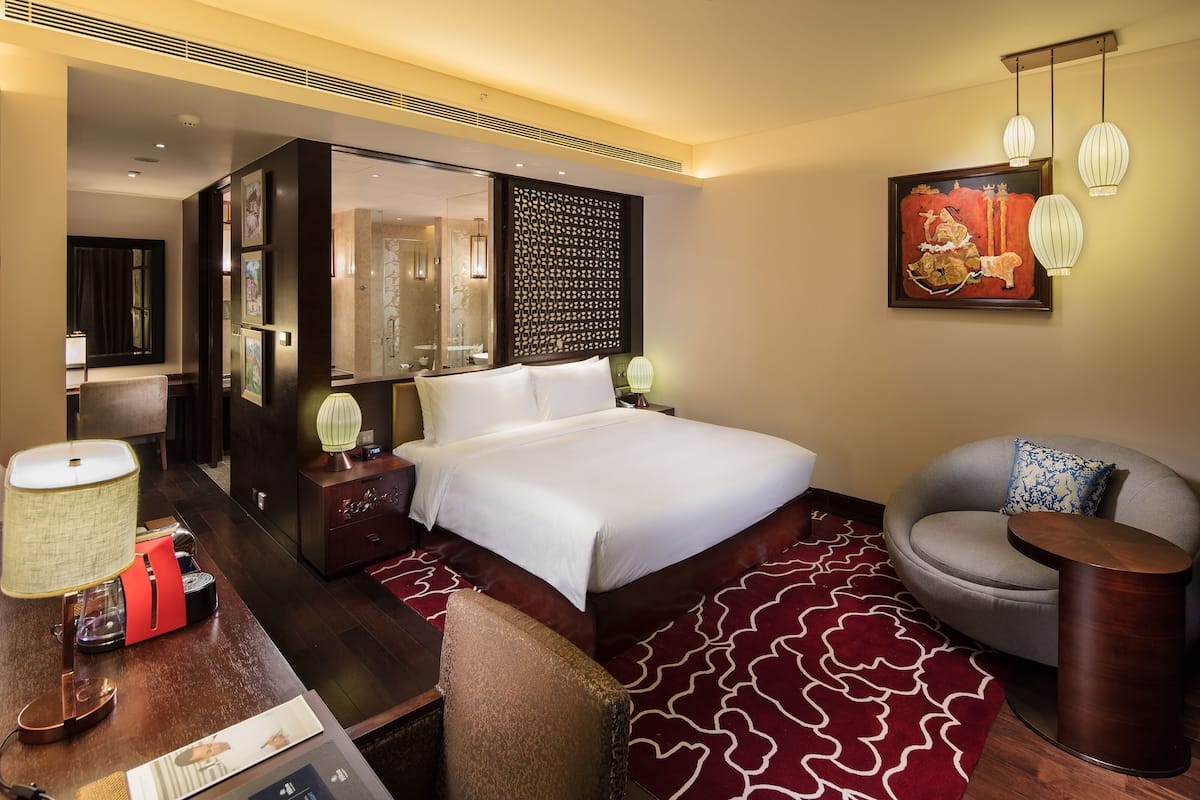 A hotel room with a large bed, a sitting area with a chair and a small table, a desk with a lamp, and a decorative wall piece.