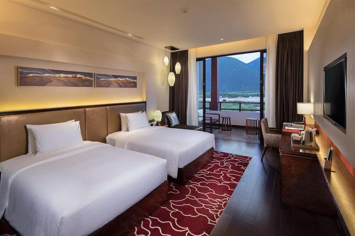 A hotel room with two beds, a TV, a desk, and a balcony with a mountain view.