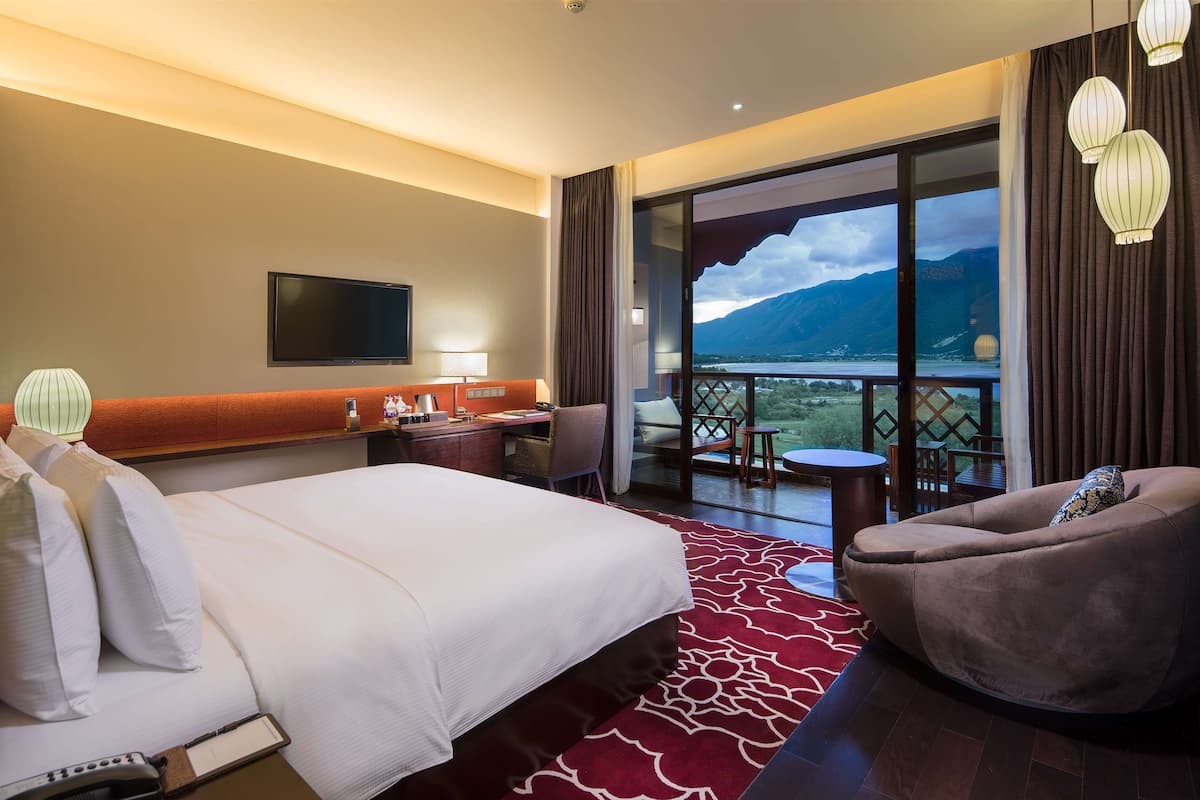 A hotel room with a bed, desk, chair, and a balcony with a view of mountains.