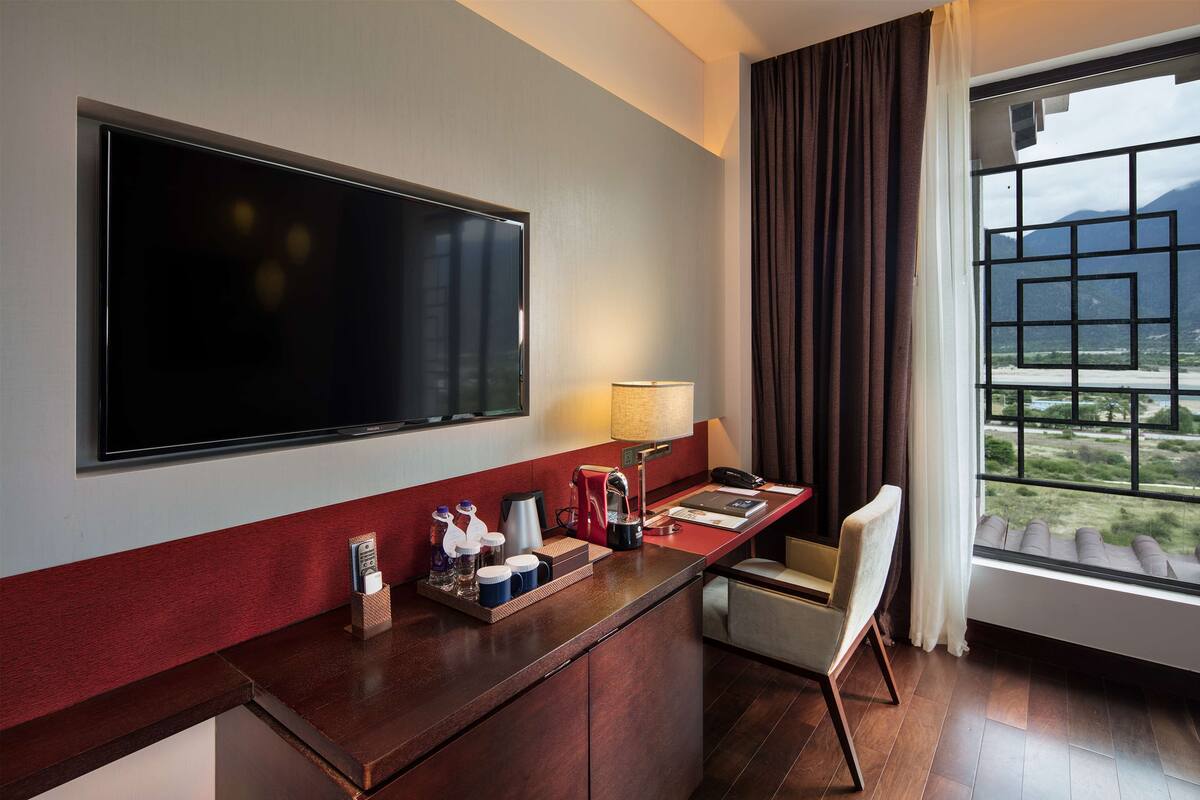 A hotel room with a large flat-screen TV mounted on the wall, a desk with a lamp, a chair, and a window with a view of mountains.