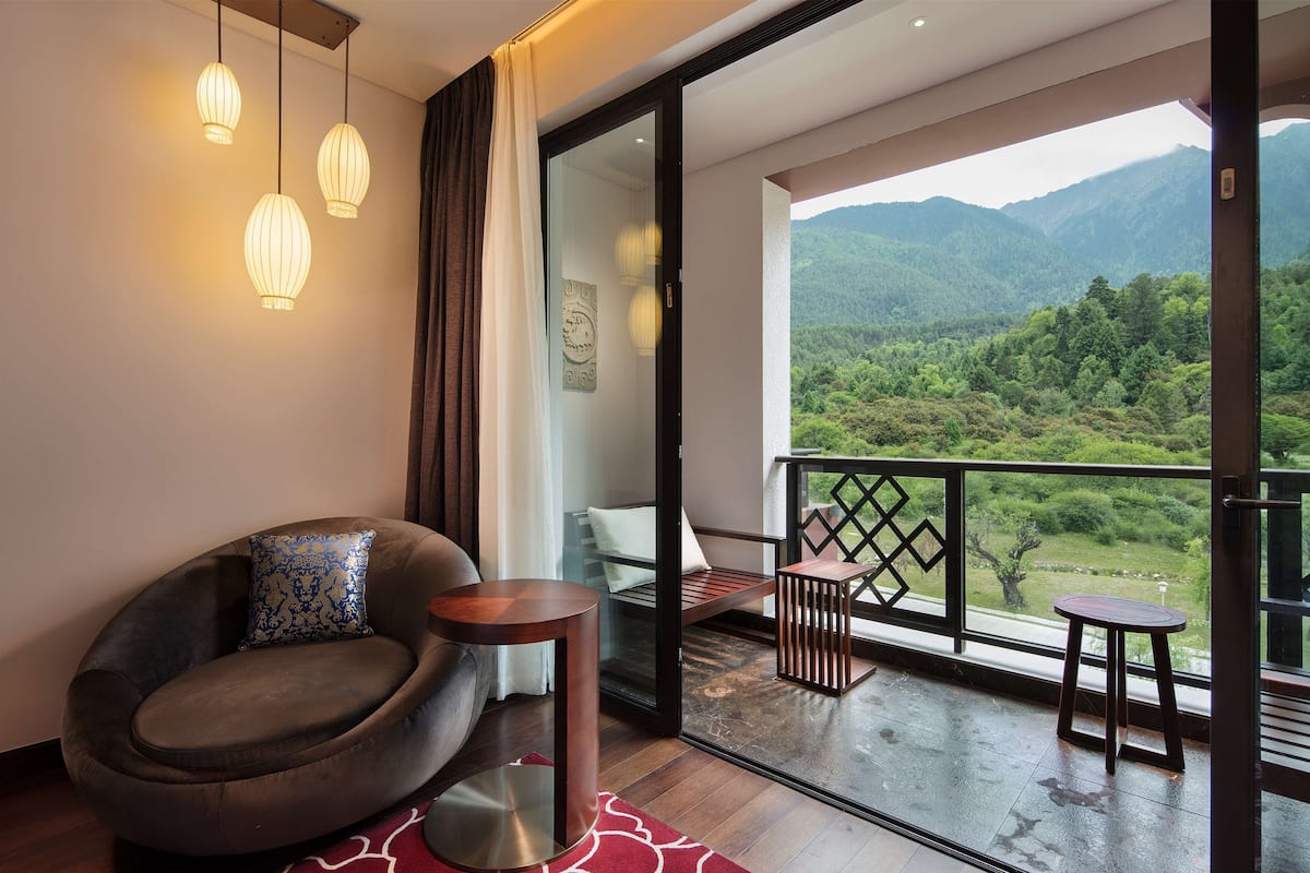 A modern room with a comfortable chair, a small table, and a balcony with a view of lush green mountains.