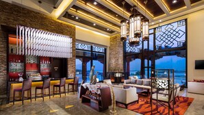 A modern bar area with a long counter, multiple bar stools, and a decorative shelf. There is a seating area with sofas and a coffee table, and large windows offering a view of the outdoors. - Hilton Linzhi Resort (Nyingtri)