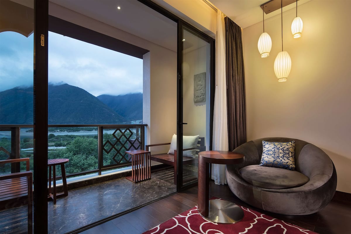 A modern room with a balcony, a round grey sofa, a small table, and a view of mountains.