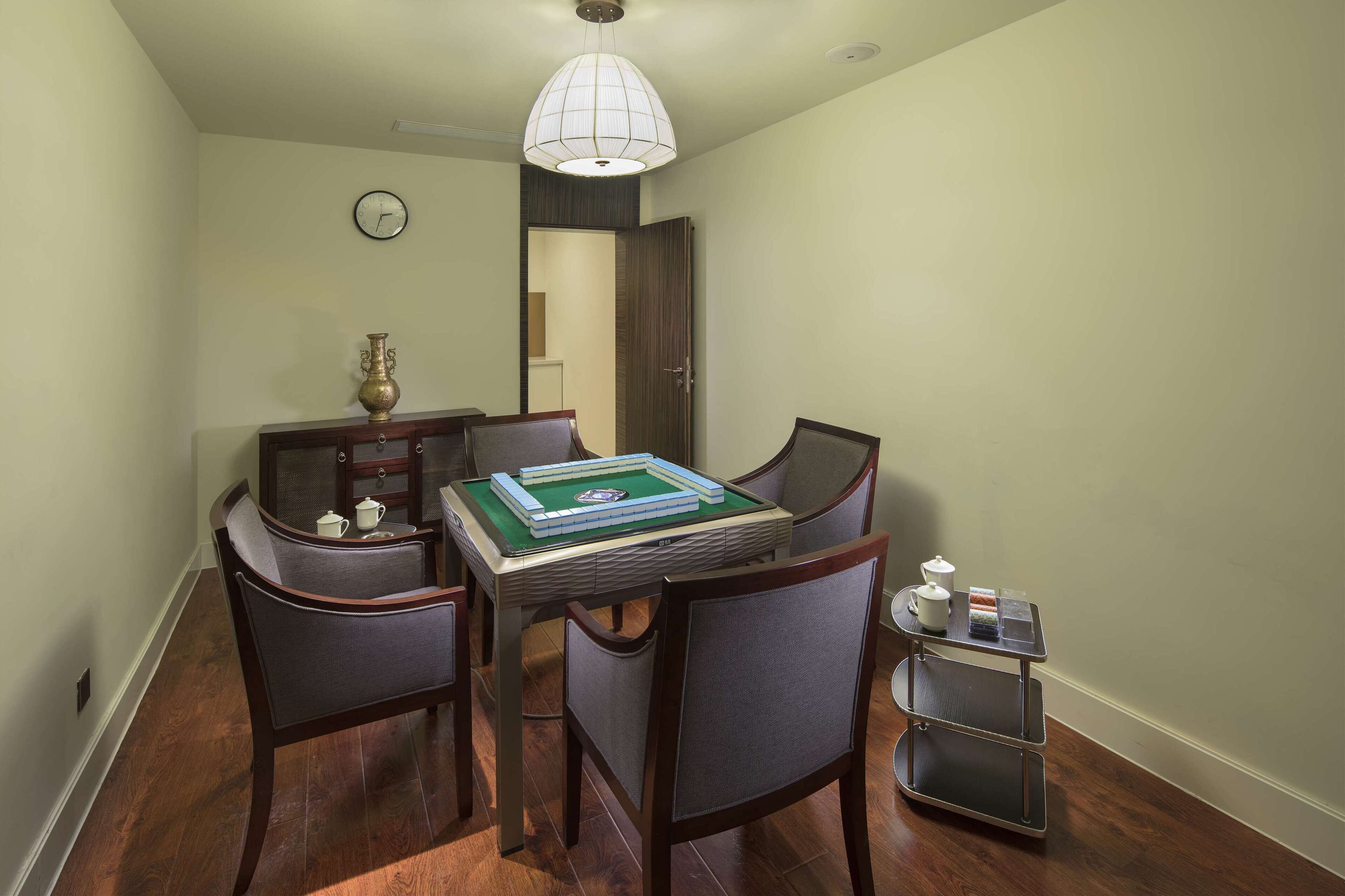 Un comedor con una mesa dispuesta para cuatro personas, sillas, una mesita auxiliar con una tetera y un reloj en la pared.