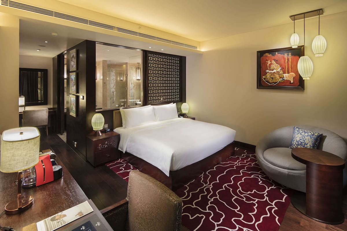 A hotel room with a large bed, a desk with a lamp, a chair, a round side table, and a decorative wall piece.