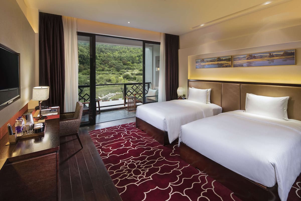 A hotel room with two beds, a TV, a desk with a lamp, and a balcony with a scenic view.