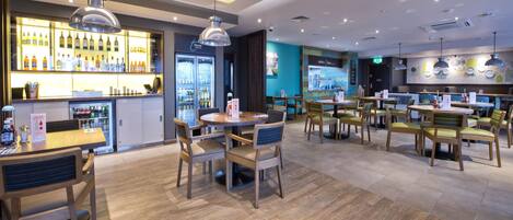 Ein modernes Restaurant mit Holztischen und -stühlen, einer Bar mit Flaschen sowie einer blauen Wand mit einer Karte.