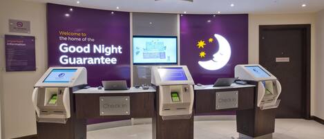 A hotel reception area with three check-in kiosks, a digital screen displaying the menu, and a sign indicating a fast track key collection for online check-ins.