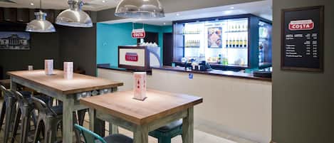 Interieur van een Costa Coffee winkel met houten tafels, teal-kleurige stoelen en een menubord.