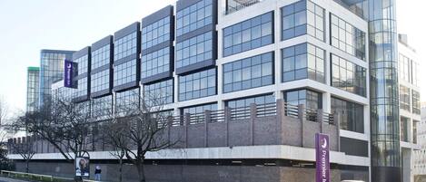 A modern hotel building with a prominent glass facade, a parking area, and a visible sign for the Premier Inn.