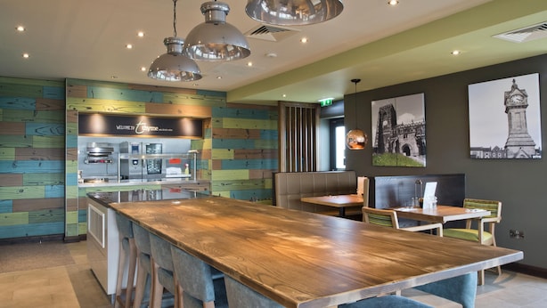 A modern restaurant interior with a long wooden table, bar stools, pendant lighting, a kitchen area, and artwork on the walls. - Premier Inn Thirsk (Thirsk)