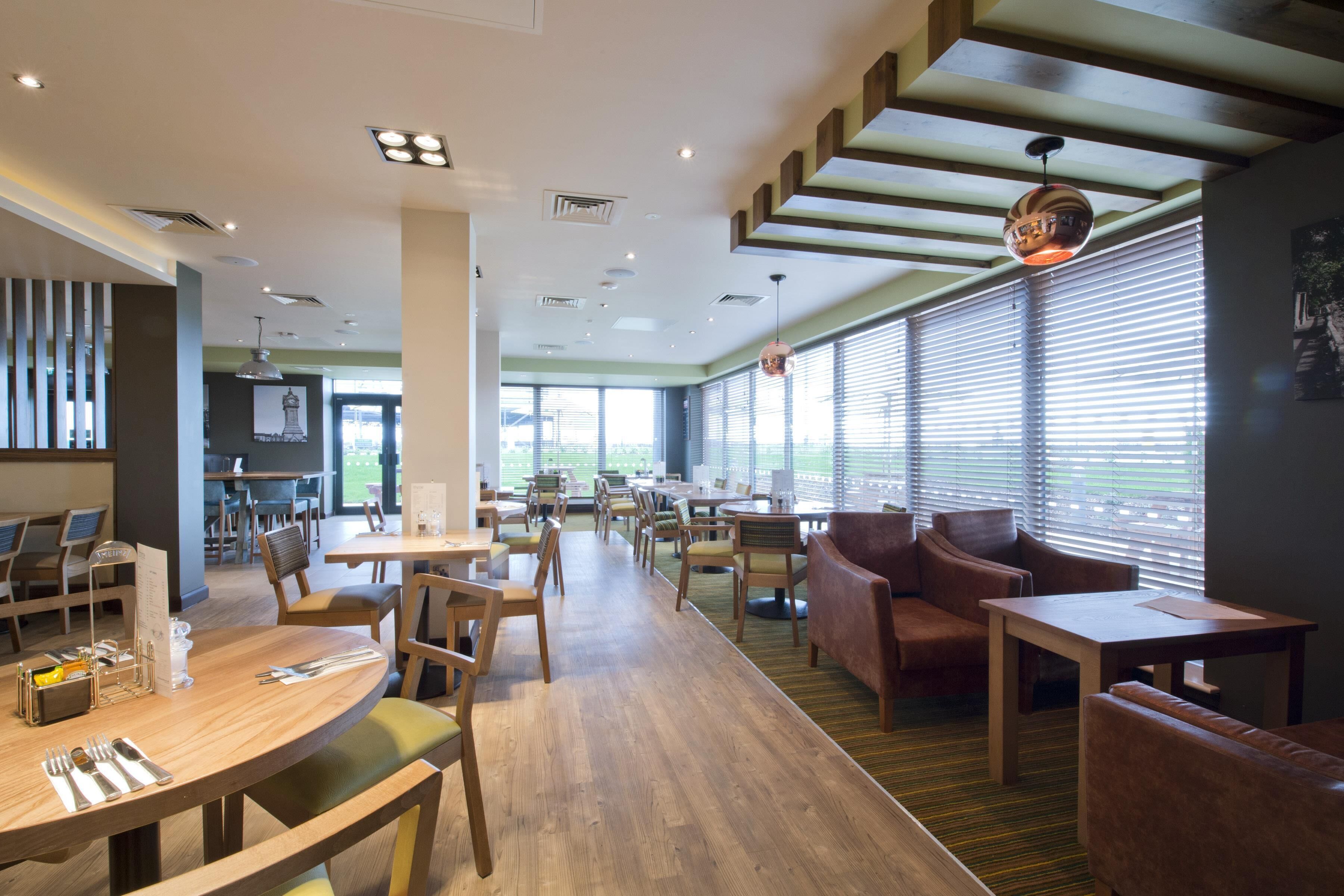 A modern restaurant with wooden tables and chairs, a bar area, and large windows with blinds.