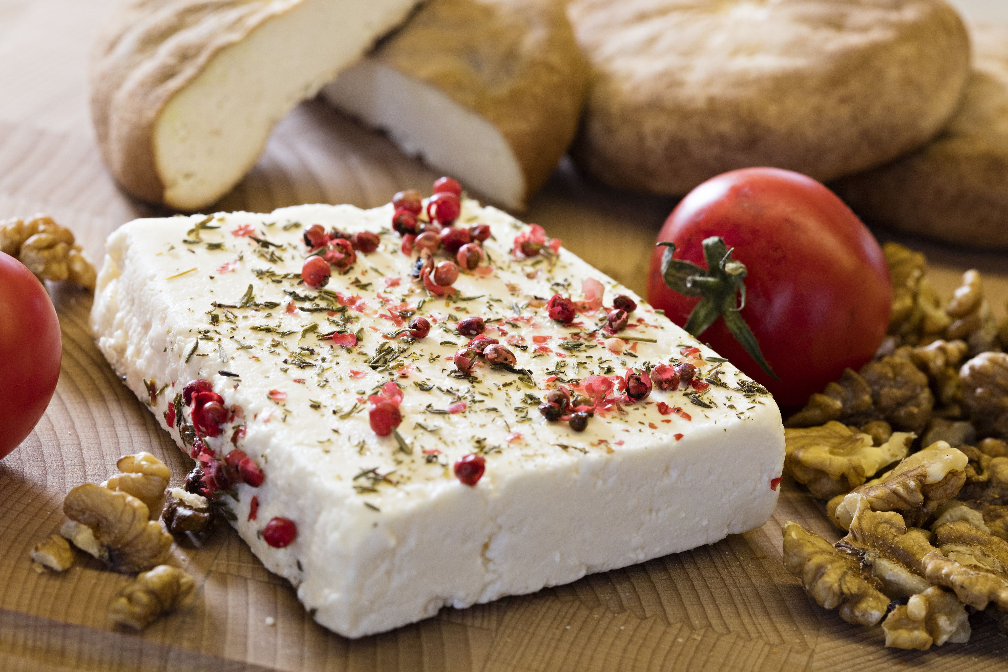 A block of cheese garnished with herbs and berries, surrounded by walnuts and tomatoes on a wooden surface.