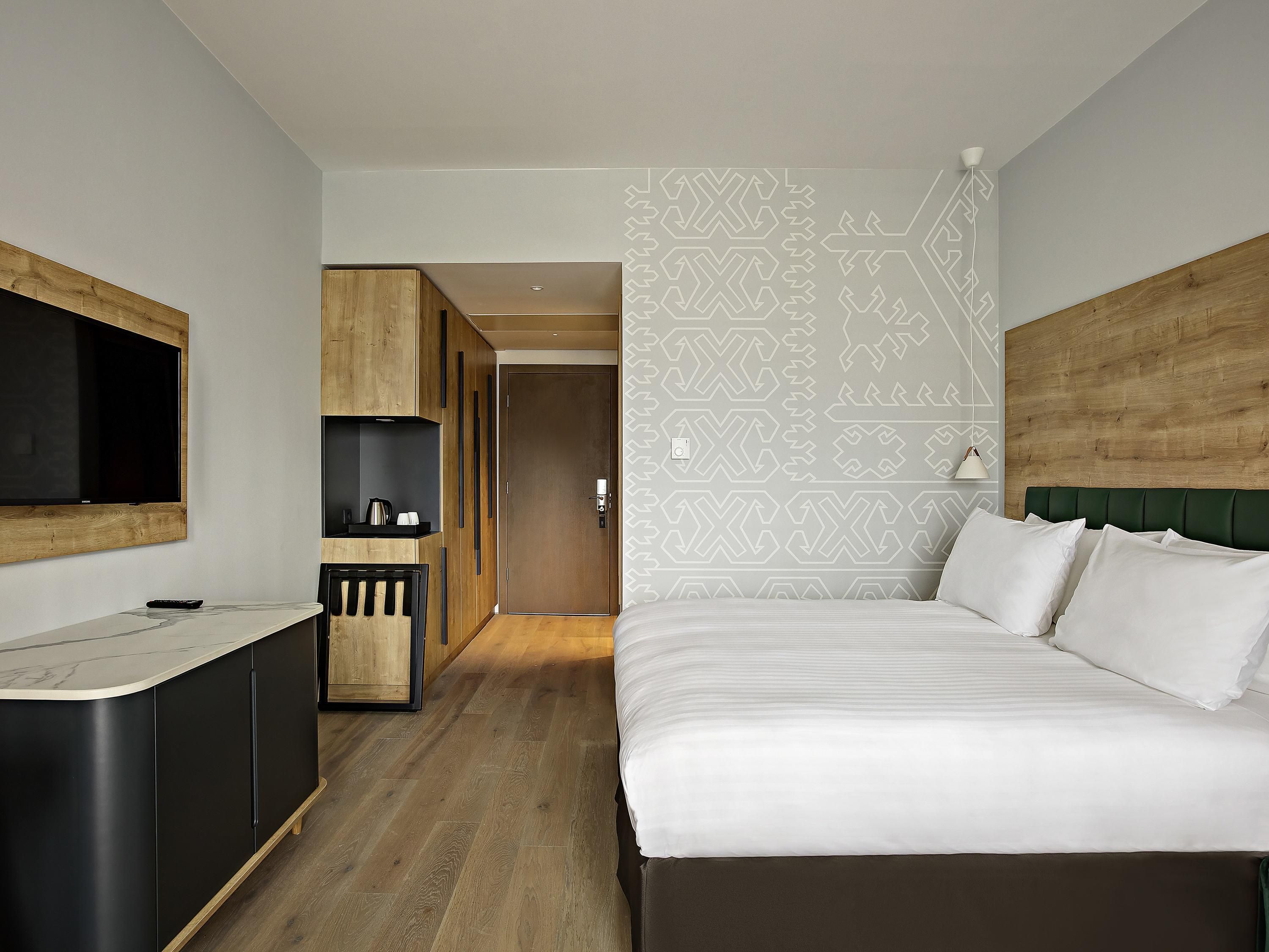 A modern hotel room with a large bed, a built-in TV, a small kitchenette, and a patterned wall.