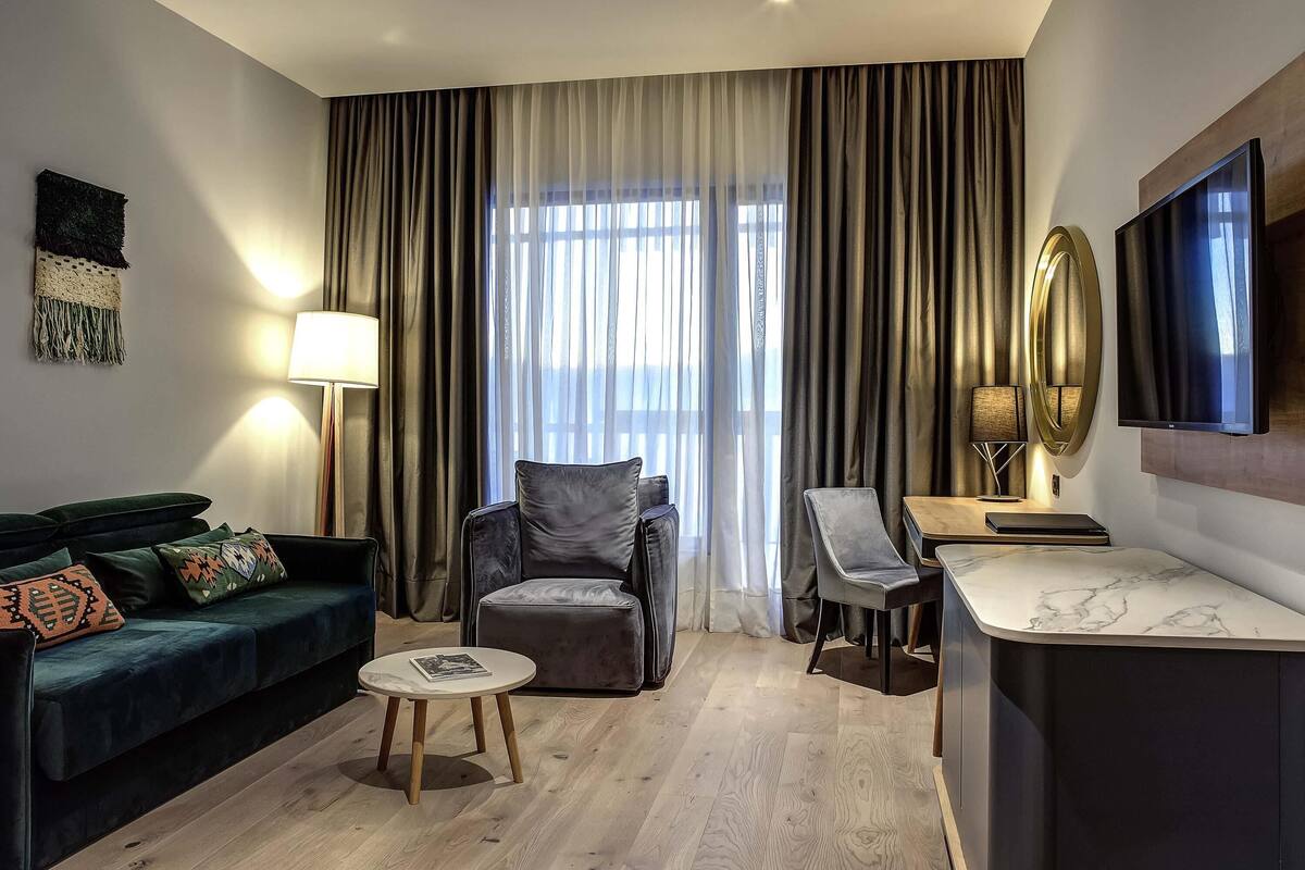 Habitación de hotel moderna con un sofá verde oscuro, una mesita redonda pequeña, un sillón gris, un escritorio de madera y un ventanal grande con cortinas.