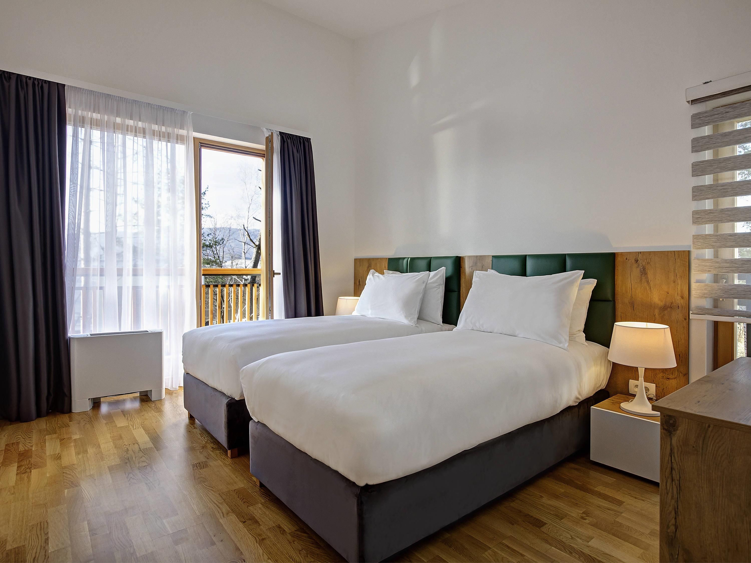 A hotel room with two beds, a wooden floor, a balcony, and a window with curtains.