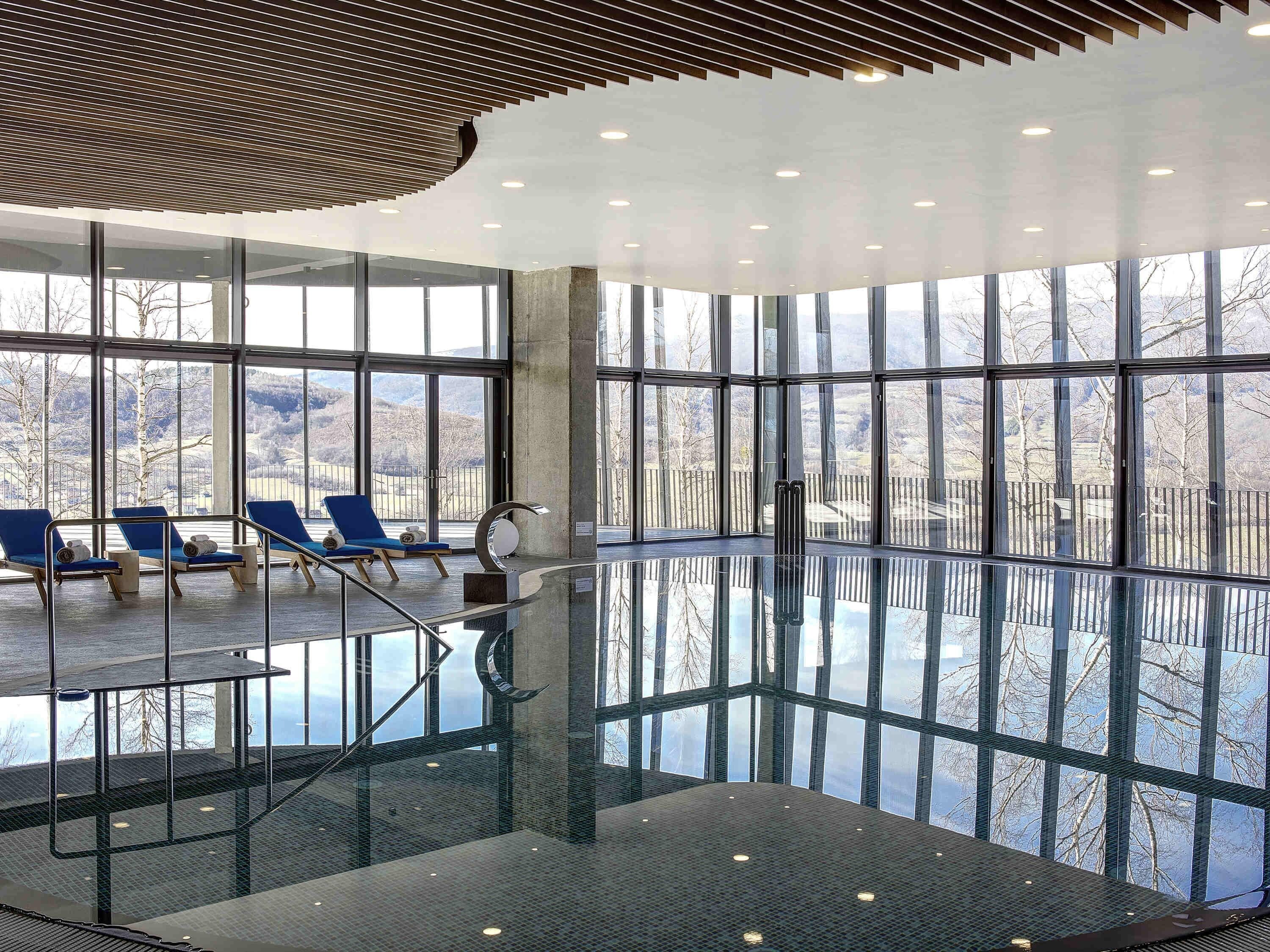 Ein modernes Hallenbad mit Glaswänden, blauen Liegestühlen und freiem Blick ins Freie.