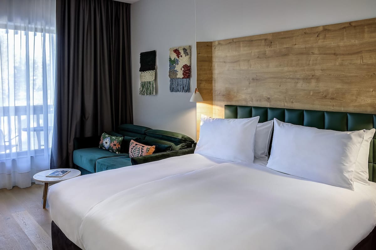 Habitación de hotel con una cama grande, un sofá verde, una mesita con un libro y una ventana con cortinas.