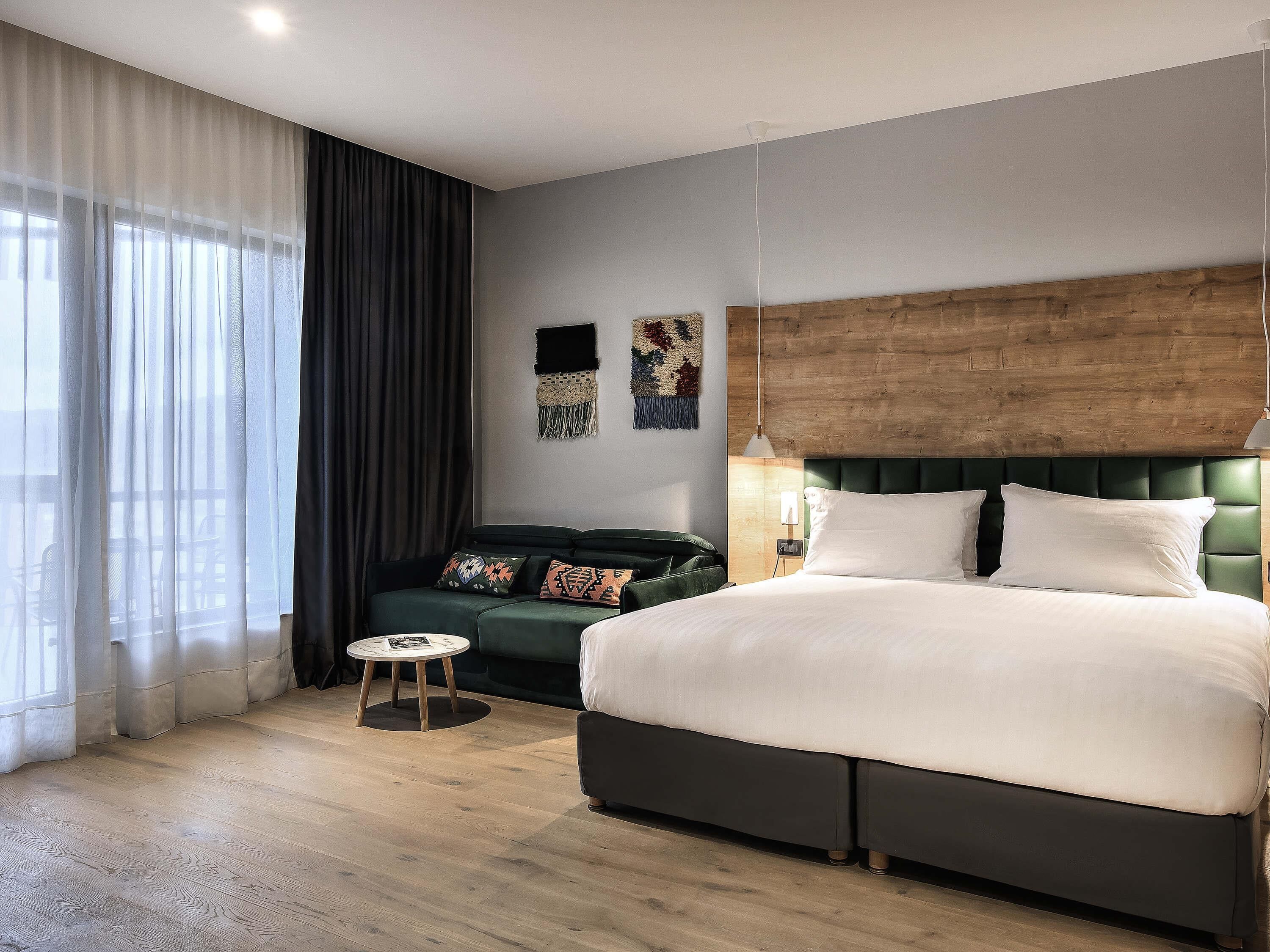 Una habitación de hotel moderna con una cama grande, un sofá verde, una mesita redonda y un cabecero de madera.