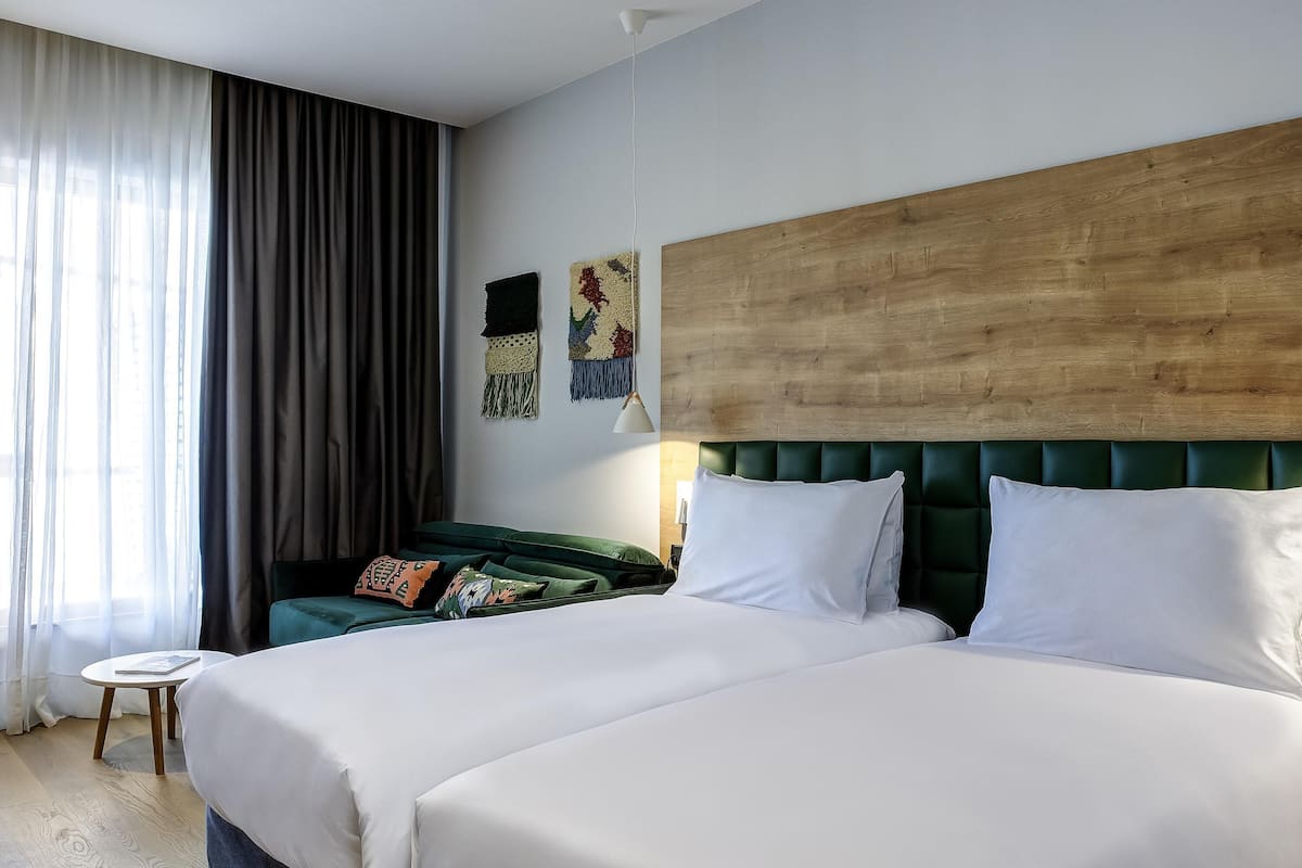 Habitación de hotel con dos camas, cabecero de madera, una mesita redonda y vistas al exterior.